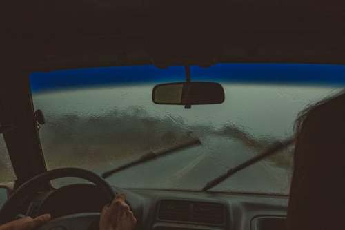 A view from inside a car with rain on the windshield and windshield wipers in motion, driving on a wet road.