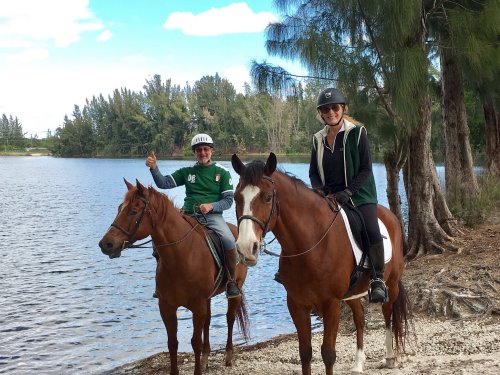 Two riders on horseback by a lake, smiling and enjoying a sunny day surrounded by trees.