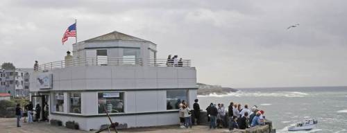 A coastal building with an American flag, surrounded by people, overlooking the ocean and a boat in the water.