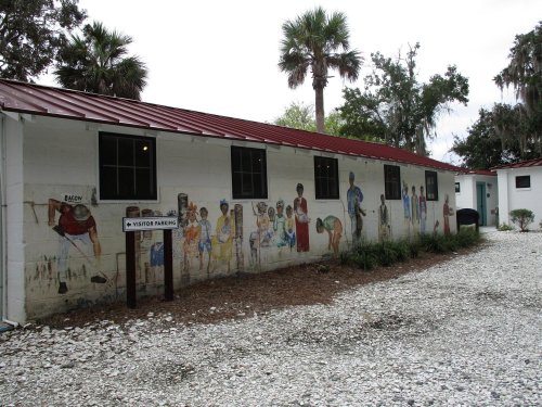 Mural-covered building exterior with palm trees and gravel parking area. Sign for visitor parking visible.