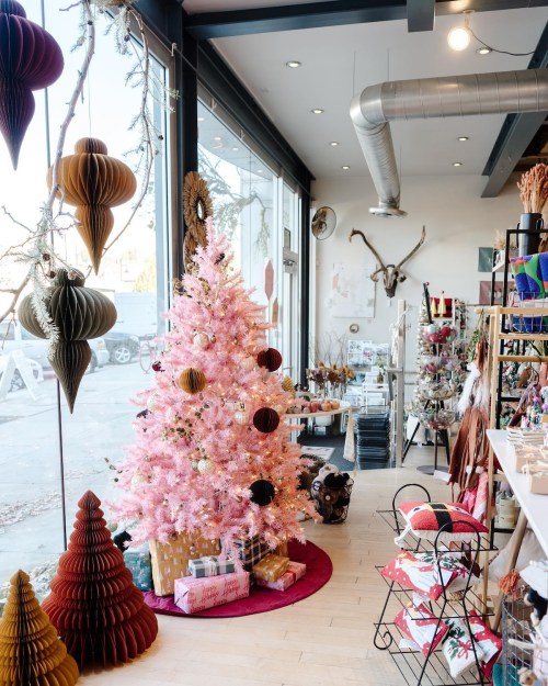 A pink Christmas tree adorned with ornaments stands in a cozy shop filled with holiday decorations and gifts.