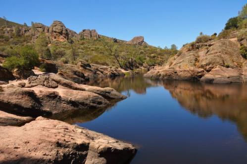 A serene landscape featuring a calm river surrounded by rocky terrain and lush greenery under a clear blue sky.