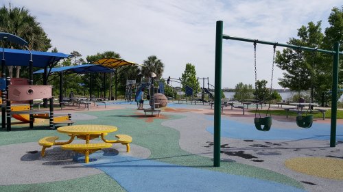 Colorful playground with swings, slides, and picnic tables, surrounded by trees and a view of water in the background.