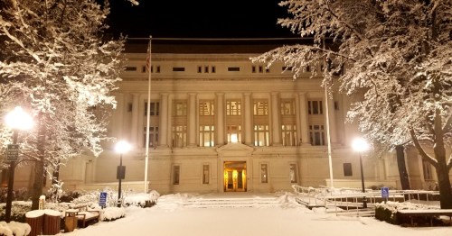 A snow-covered building at night, illuminated by street lamps, surrounded by trees and a quiet, wintry landscape.