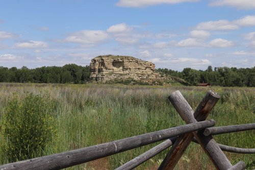 A rocky outcrop rises above a grassy field, framed by a wooden fence under a blue sky with fluffy clouds.