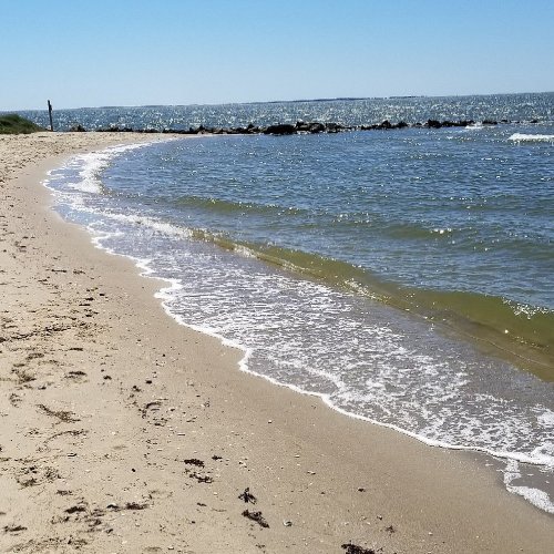 A sandy beach curves along a calm shoreline with gentle waves lapping at the shore under a clear blue sky.