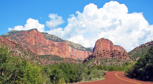 Scenic view of red rock formations and mountains under a blue sky with fluffy white clouds, alongside a winding road.