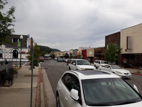 A street view of a small town with parked cars, shops, and cloudy skies in the background.