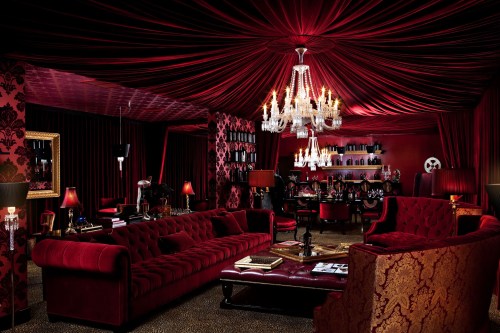 Luxurious room with red drapery, plush sofas, a chandelier, and a stylish bar area, creating an elegant atmosphere.