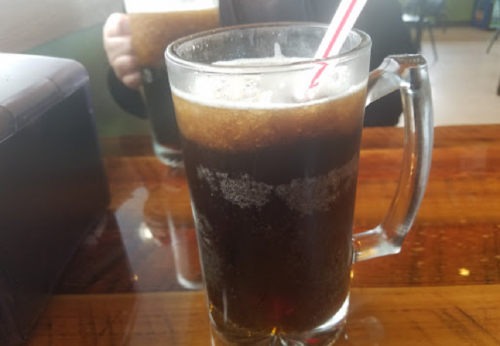 Two frosty mugs of dark soda with straws, sitting on a wooden table.