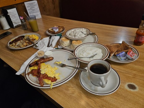 A table with plates of breakfast food, including eggs, bacon, sausage, grits, biscuits, toast, and a cup of coffee.