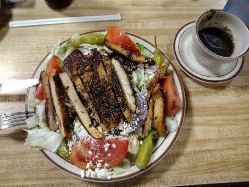 A plate of salad topped with grilled chicken, tomatoes, peppers, and feta cheese, with a cup of dressing on the side.