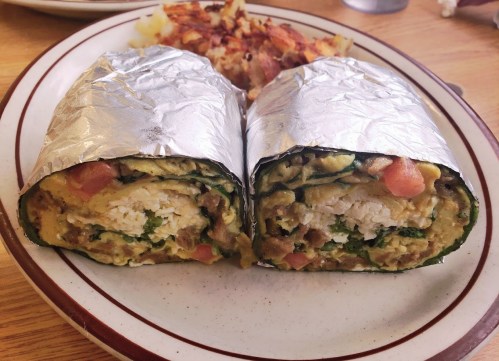 A cut burrito wrapped in foil, filled with eggs, cheese, and vegetables, served with a side of hash browns.