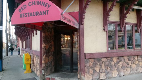 Exterior of Red Chimney Restaurant with a red awning and stone facade, featuring a glass entrance and nearby sidewalk.