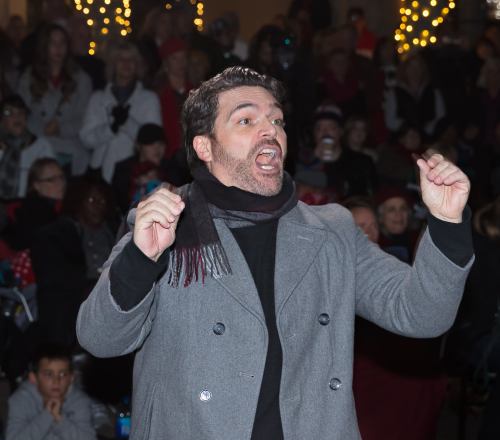 A man in a gray coat passionately gestures while speaking to a crowd during an outdoor event.