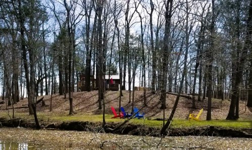 A serene lakeside scene with colorful chairs on the shore, surrounded by trees and a cabin in the background.