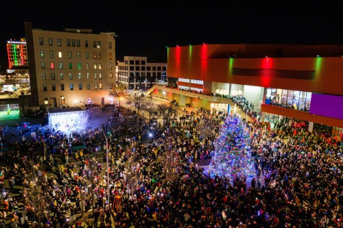 A large crowd gathers around a brightly lit Christmas tree at night, with festive lights on nearby buildings.