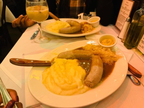 A plate of sausages with mashed potatoes, gravy, and sauerkraut, accompanied by a drink in a glass.