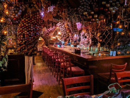 A cozy bar adorned with colorful decorations and hanging ornaments, featuring wooden chairs and a well-stocked counter.