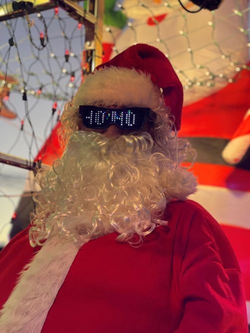A festive Santa Claus with a fluffy white beard and sunglasses, surrounded by holiday decorations and lights.