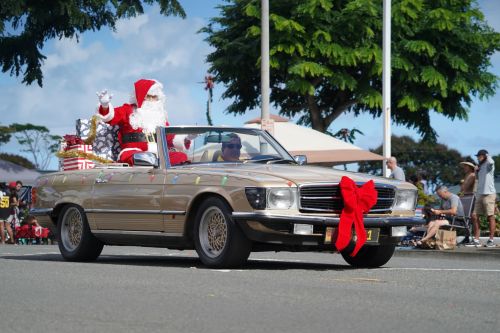 A festive convertible car with Santa and gifts, decorated with a large red bow, driving in a parade.
