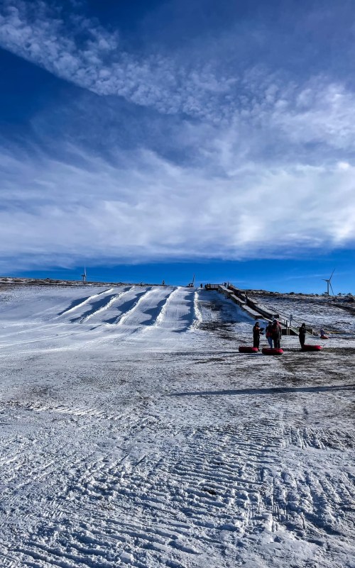 A snowy hill with several sledding lanes and people preparing to slide down under a blue sky with clouds.
