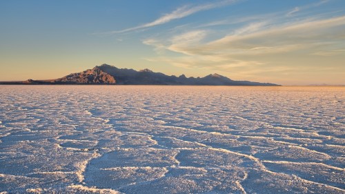 A vast salt flat stretches towards distant mountains under a clear sky, with intricate patterns on the ground.