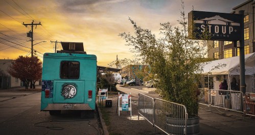 A turquoise food truck parked on a street at sunset, with a sign for "Stoup Brewing" in the background.