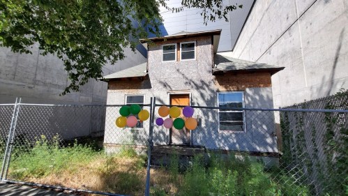 Abandoned house with a fenced yard, featuring colorful balloons on the front, surrounded by concrete walls.