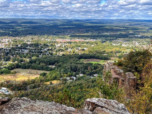 A panoramic view of a lush valley with towns, fields, and rolling hills under a partly cloudy sky.