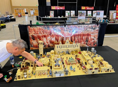 A person adjusts a large LEGO display featuring intricate buildings and scenes, set against a backdrop of red rock formations.