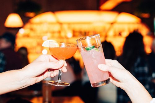 Two hands clink glasses in a bar, one holding a cocktail and the other a refreshing pink drink with mint.