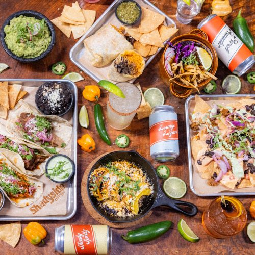 A vibrant spread of tacos, nachos, guacamole, drinks, and sides on a wooden table, showcasing a festive meal.