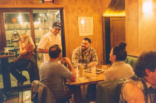 A lively restaurant scene with diners chatting and a server interacting with guests at a wooden table.
