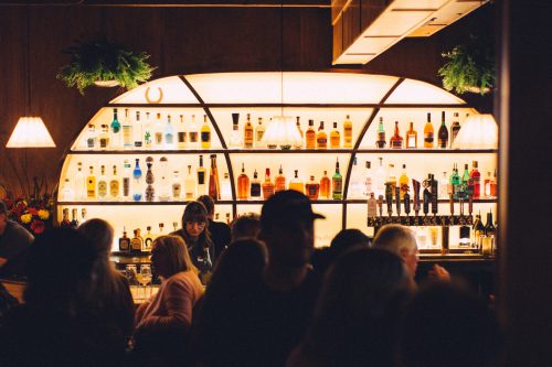 A dimly lit bar with a backlit shelf of colorful liquor bottles and a bartender serving drinks to a crowd.