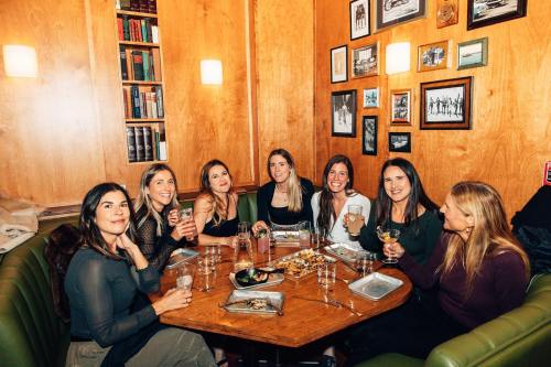 A group of seven women enjoying drinks and food at a cozy restaurant table, smiling and chatting together.