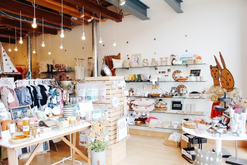 A cozy boutique featuring clothing racks, shelves of home goods, and decorative items under warm hanging lights.