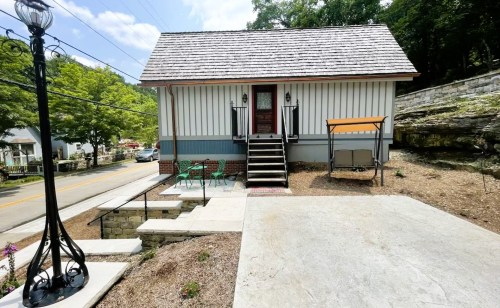 A small, charming house with a porch, steps, and outdoor seating, surrounded by greenery and a quiet street.