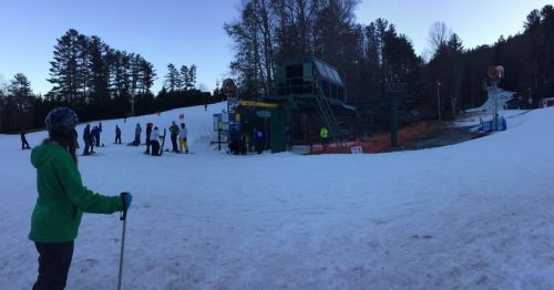 A ski resort scene with people waiting near a ski lift on a snowy slope surrounded by trees at dusk.