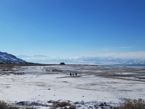 A snowy landscape with people walking near a frozen lake, surrounded by mountains under a clear blue sky.