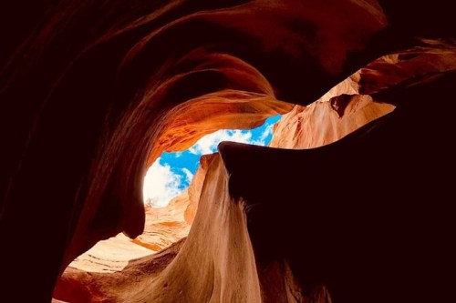 A view of a canyon with swirling rock formations and a bright blue sky peeking through the opening above.