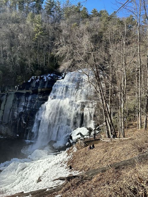 A waterfall cascades over rocks, surrounded by trees, with ice at the base and a small group of people nearby.