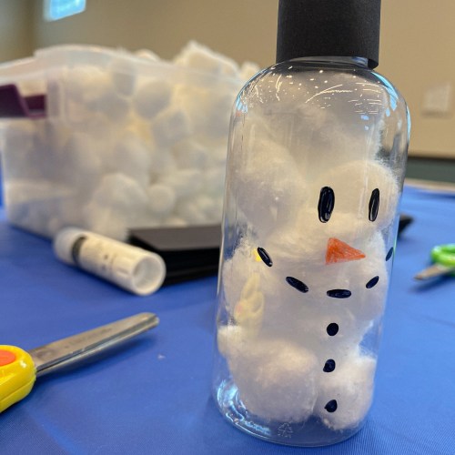 A clear bottle filled with cotton balls shaped like a snowman, featuring a black cap, orange carrot nose, and button details.