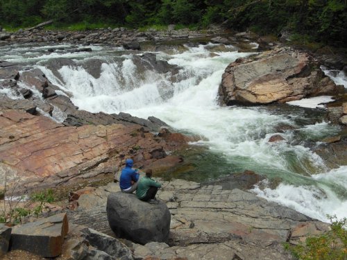 Two people sit on a rock by a rushing river, surrounded by lush greenery and rocky terrain.