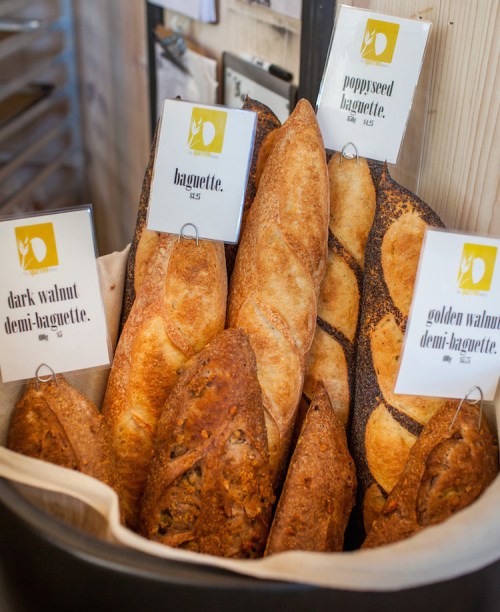 A variety of artisanal breads displayed in a basket, including dark walnut, poppyseed, and golden walnut demi-baguettes.