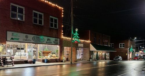 A festive street scene at night, featuring decorated storefronts and a Christmas tree, with wet pavement reflecting lights.