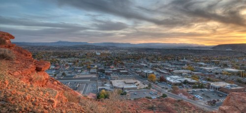 A panoramic view of a city at sunset, surrounded by rocky terrain and distant mountains under a colorful sky.