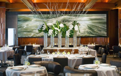 Elegant restaurant interior with round tables, floral centerpieces, and a large ocean-themed mural on the wall.
