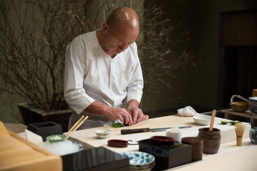 A chef in a white uniform skillfully prepares sushi at a wooden counter, surrounded by traditional Japanese utensils.