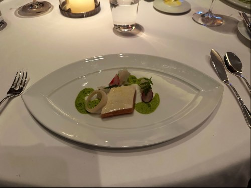 A plated dish featuring fish, green sauce, and garnishes, set on a white tablecloth with cutlery and glassware.